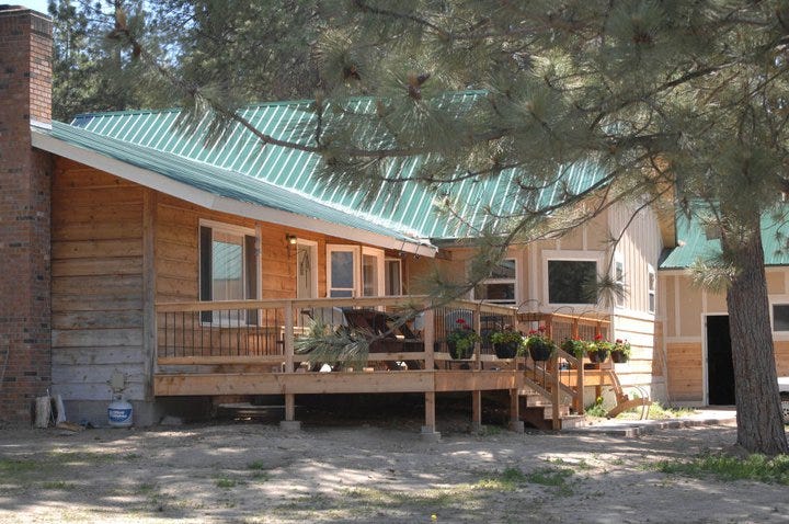 a spacious wooden deck decorated with baskets of flowers on the front of a large home surrounded by pine trees