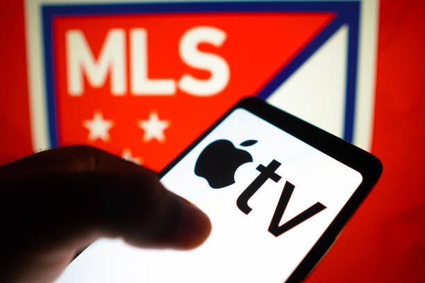 In this photo illustration, the Apple TV logo is displayed on a smartphone screen and Major League Soccer logo in the background. MLS and Leagues Cup...