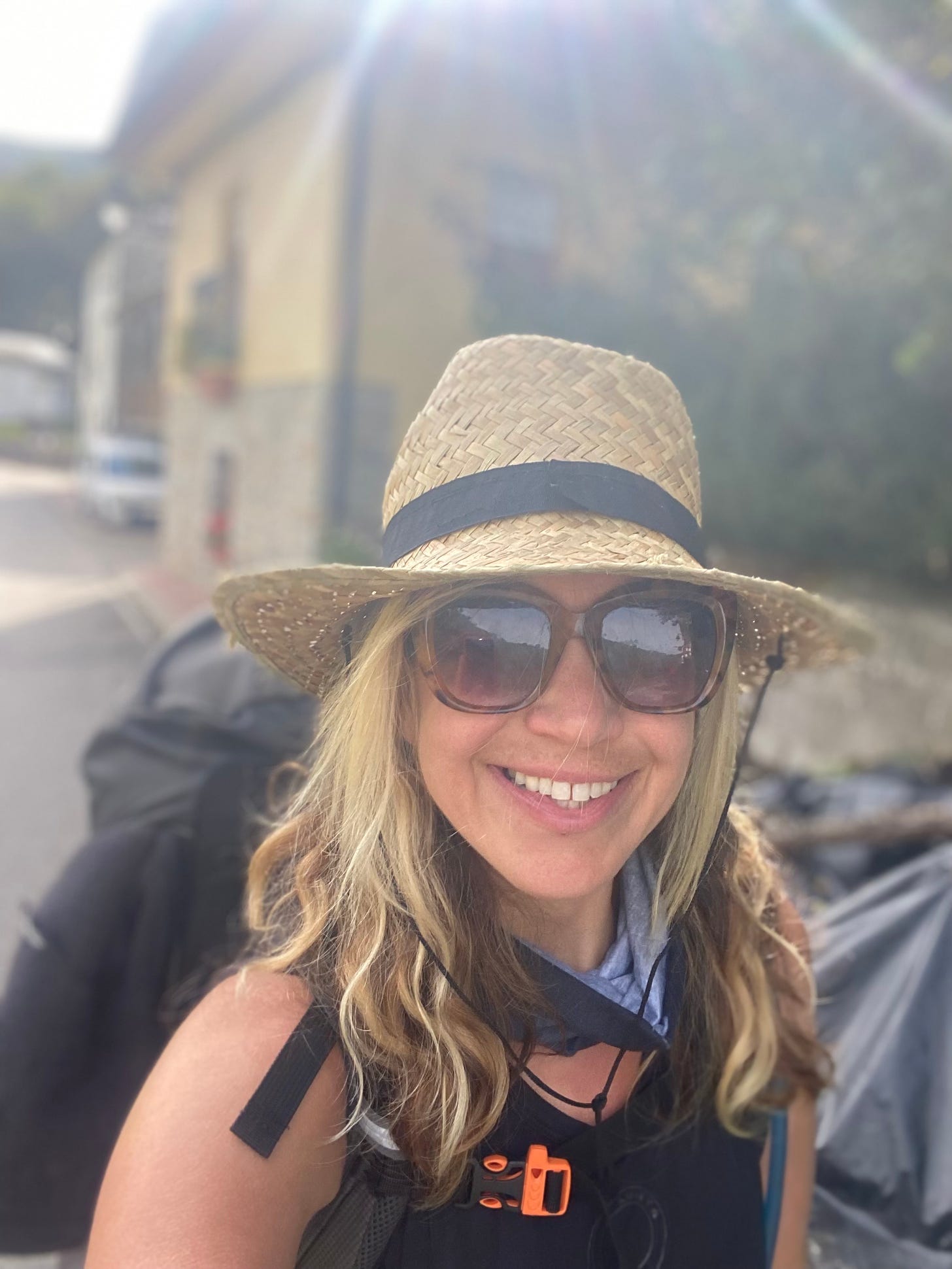 A woman in a hat and sunglasses, wearing a backpack smiles, pausing on her walk of the Camino de Santiago.