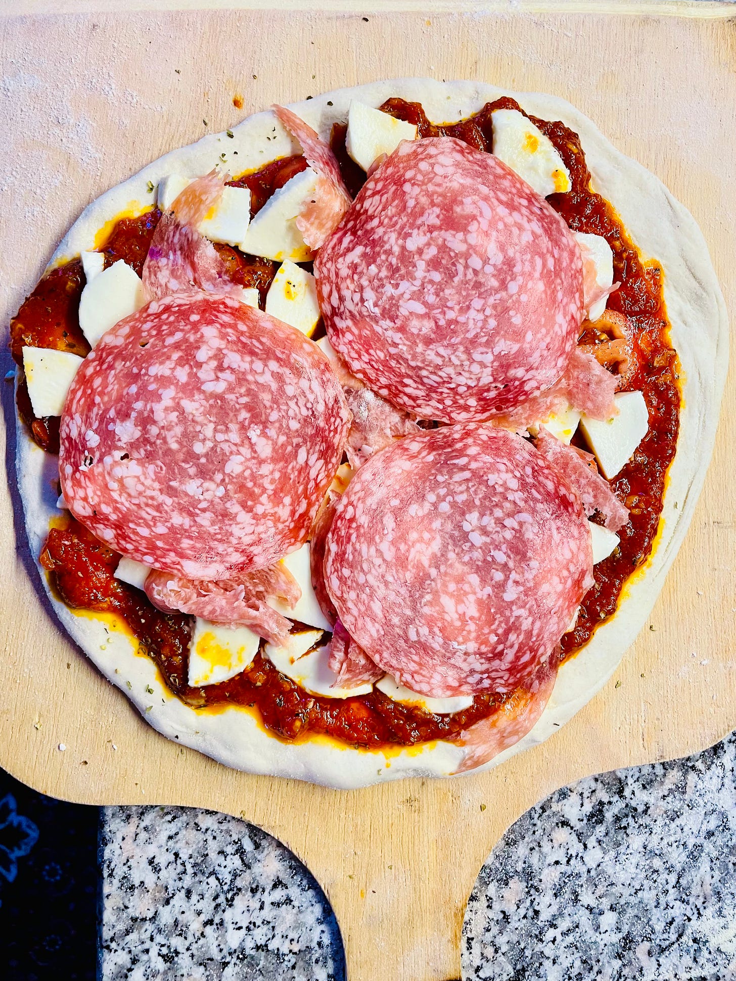 An overhead view of an unbaked pizza on a wooden peel, topped with a thick layer of tomato sauce, chunks of fresh mozzarella, and three large slices of Milano salami placed evenly across the surface. The raw dough edges are visible, dusted with flour, and the pizza is prepped for the oven. An overhead view of an unbaked pizza on a wooden peel, topped with a thick layer of tomato sauce, chunks of fresh mozzarella, and three large slices of Milano salami placed evenly across the surface. The raw dough edges are visible, dusted with flour, and the pizza is prepped for the oven.