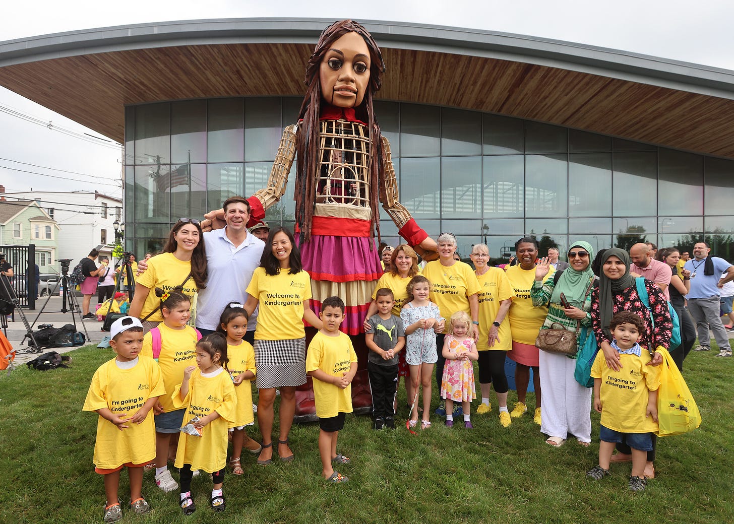 The Countdown to Kindergarten organizers, local electeds, and incoming Kindergarteners smile for a picture with the 12-foot puppet, Little Amal, outside the East Boston library