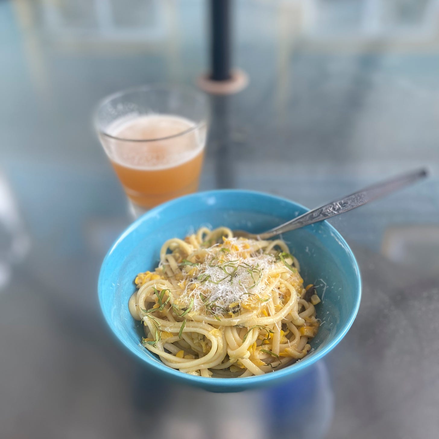 A blue bowl of linguine in the corn, olive, & zucchini mixture described above, topped with parmesan and basil chiffonade. A fork sticks out of the back of the bowl, and a glass of beer is on the table in the background.