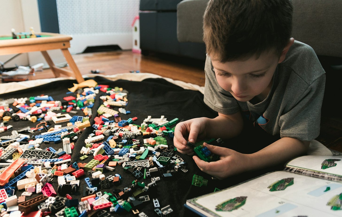 A young boy lying on a living room carpet playing lego and looking at a lego book.