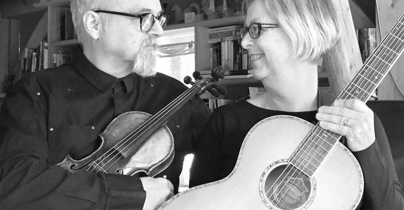 Two sixty-something musicians look at each other while holding a guitar and fiddle.
