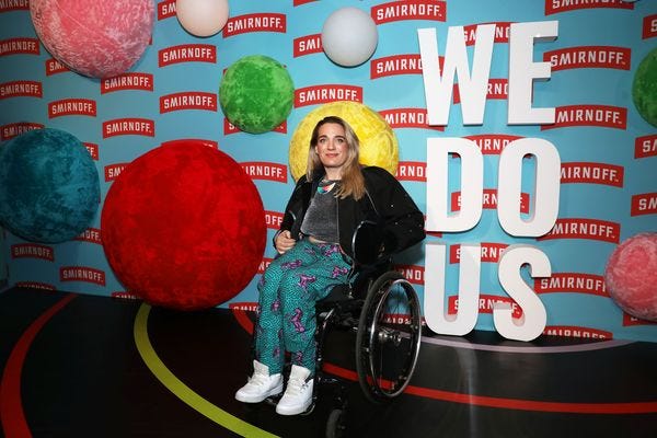 Wearing pony printed trousers, a silver top, black jacket and white trainers, I pose in my wheelchair in front of Smifnoff branding and large lettering that reads WE DO US 
