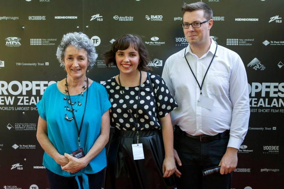 A photograph in front of a press backdrop with the logo for Tropfest New Zealand and their logos, in front of it are three people - one is Louise's mum on the left, Louise in the middle, and Blair on the right. They're all smiling at the camera and wearing Tropfest lanyards. 