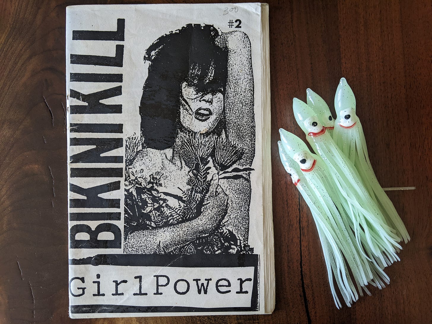 On a walnut wood table is an old zine with the words 'BIKINI KILL GirlPower' #2 next to a grainy photo of a woman on the cover. Next to the zine on the table are 5 glow-in-the-dark fishing lure squid because why not.