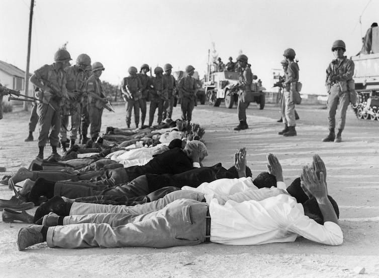Men lying down on the ground with their hands behind their heads, overseen by armed soldiers.