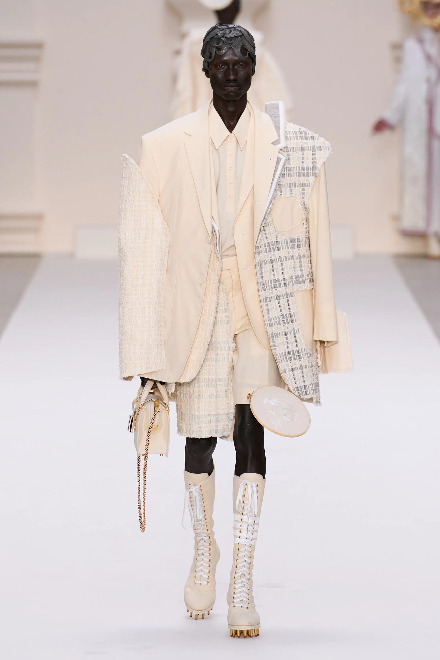 A dark-skinned male model walks down a stark white runway wearing a suit that appears spliced together from muslin and tweed.