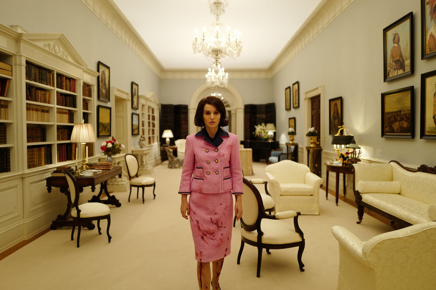 A woman walks down a hallway wearing a pink suit. The suit is covered in blood. There are bookcases and paintings on the walls.
