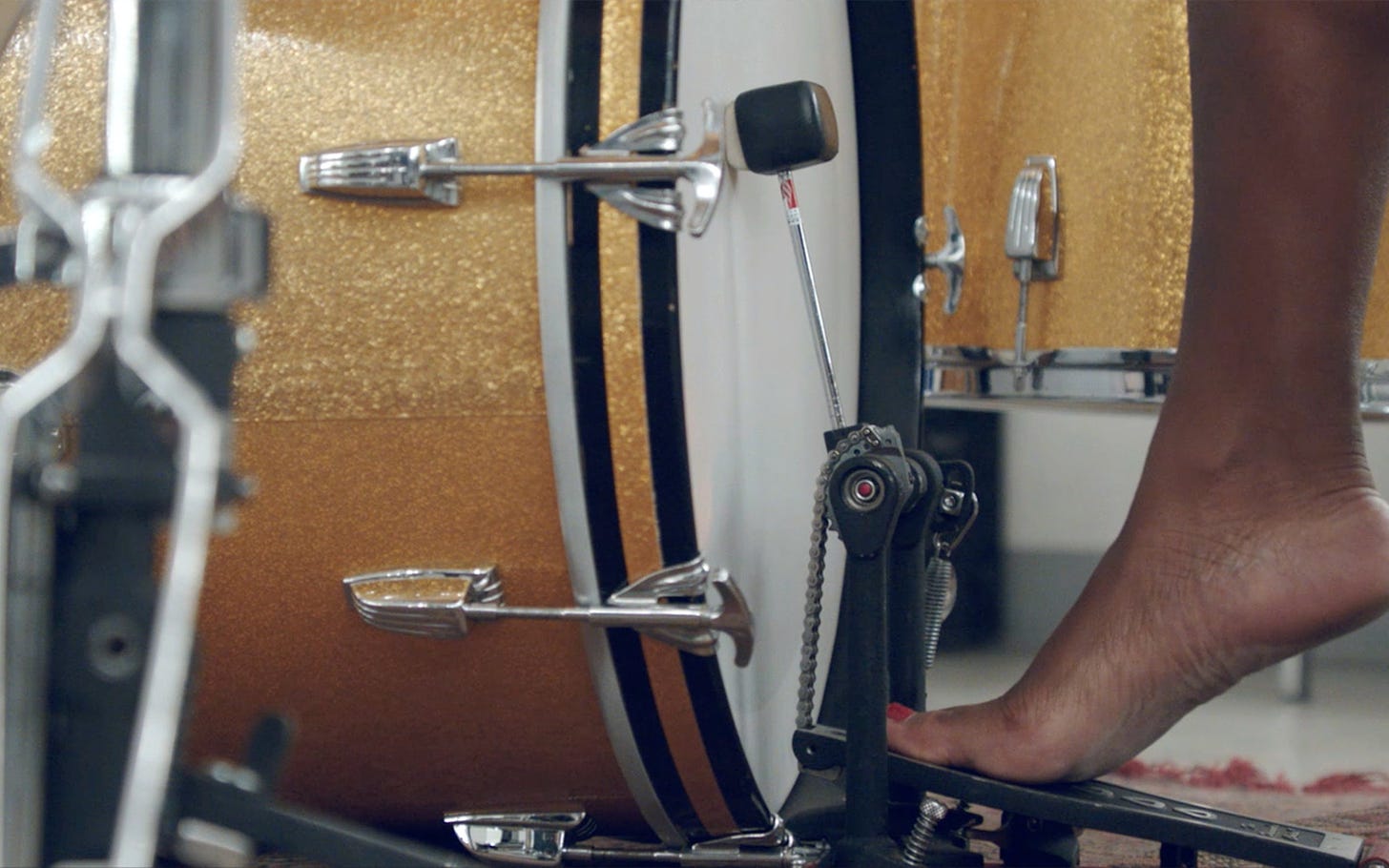 A close up shot of a medium-dark skin tone bare foot stepping on a kick drum pedal within a sparkly gold drum set.