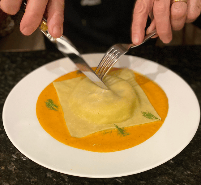 One giant ravioli in an orange coloured sauce in the center of a white plate lightly garnished with dill, hands with a fork and knife are about to slice it open.