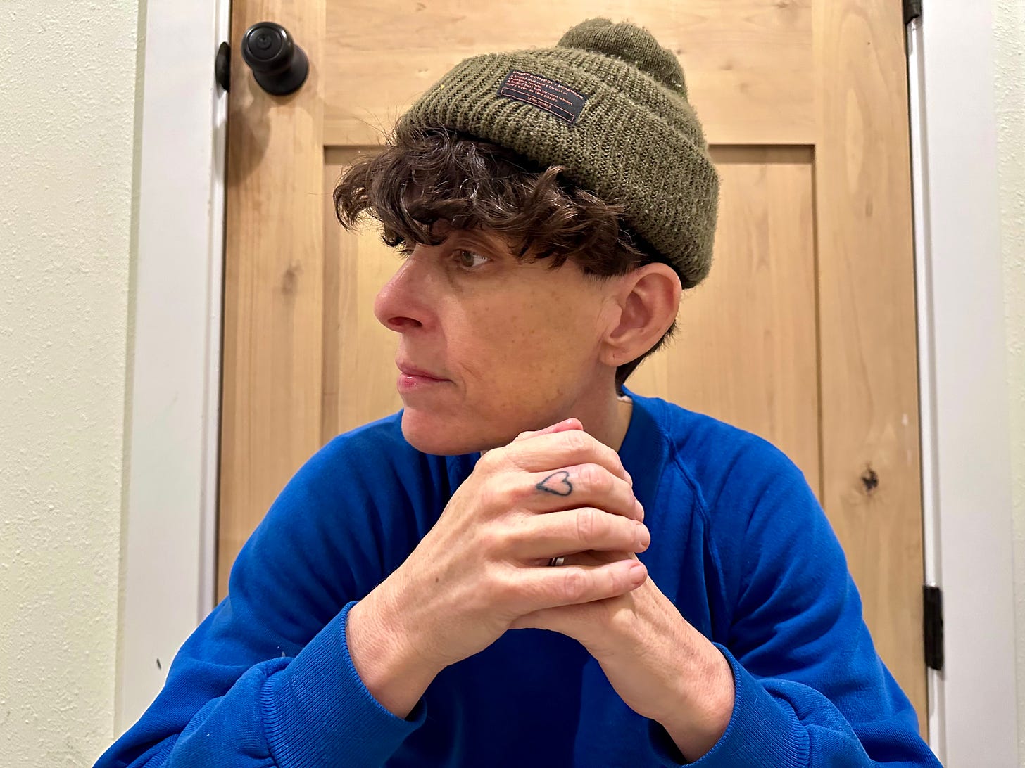 Andrea Gibson with short, curly dark hair wearing an olive-green knit beanie and a royal blue sweater. They are seated indoors against a wooden door with white walls, resting their clasped hands under their chin. A small heart tattoo is visible on their left ring finger. Their gaze is directed to the left, giving a thoughtful and introspective expression.