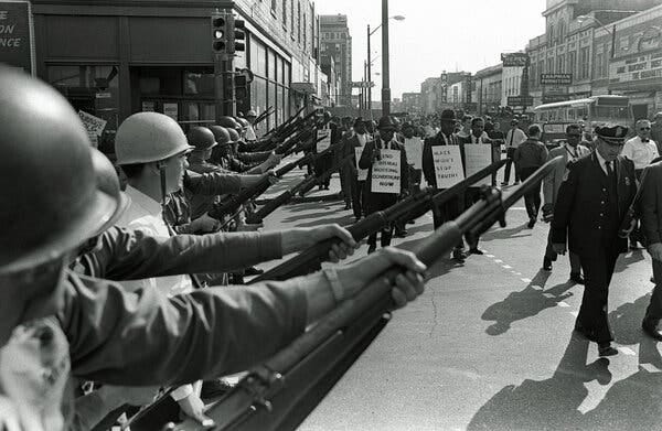 A black and white photo of people marching in a street lined with men in uniform and hardhats holding bayonets in front of them.