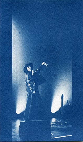Photo cyanotype de la chanteuse Fishbach sur scène. Elle porte un chapeau et des vêtements drapés. Elle porte le micro à la bouche de la main droite et sa main gauche est en l'air.