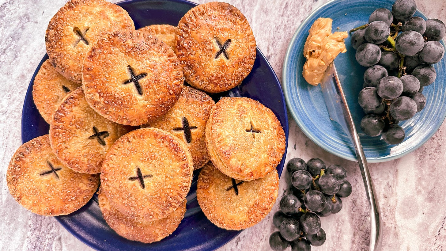 Peanut butter and concord grape jelly hand pies on a blue plate sitting next to a bunch of concord grapes and a knife covered in peanut butter.