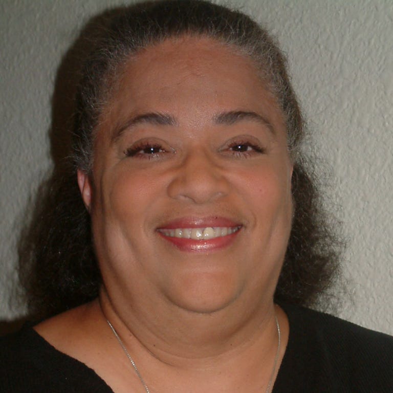 A Black woman smiles at the camera, her hair pulled back, wearing a black shirt and a thin silver necklace.