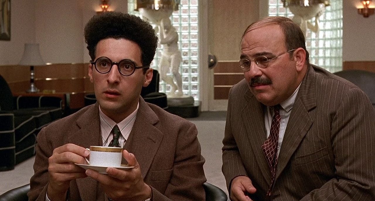 Still from Barton Fink: Barton sits in the producer's office, holding a cup of coffee on a saucer with a stunned look on his face. The producer's assistant sits at his shoulder.