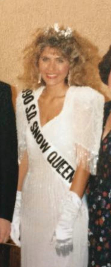 White woman with big blonde hair smiles at the camera. She wears a sparkly tiara and earrings. A sash covering her white dress reads, 1990 S.D. Snow Queen.