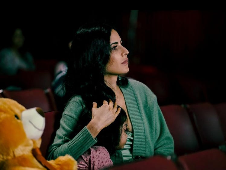 A still from Hindi film, ‘Merry Christmas’. Maria (Katrina Kaif) in a dull green dress, side-hugging a little girl with a huge teddy on the side. They are sitting in what looks like an auditorium or a theatre. 