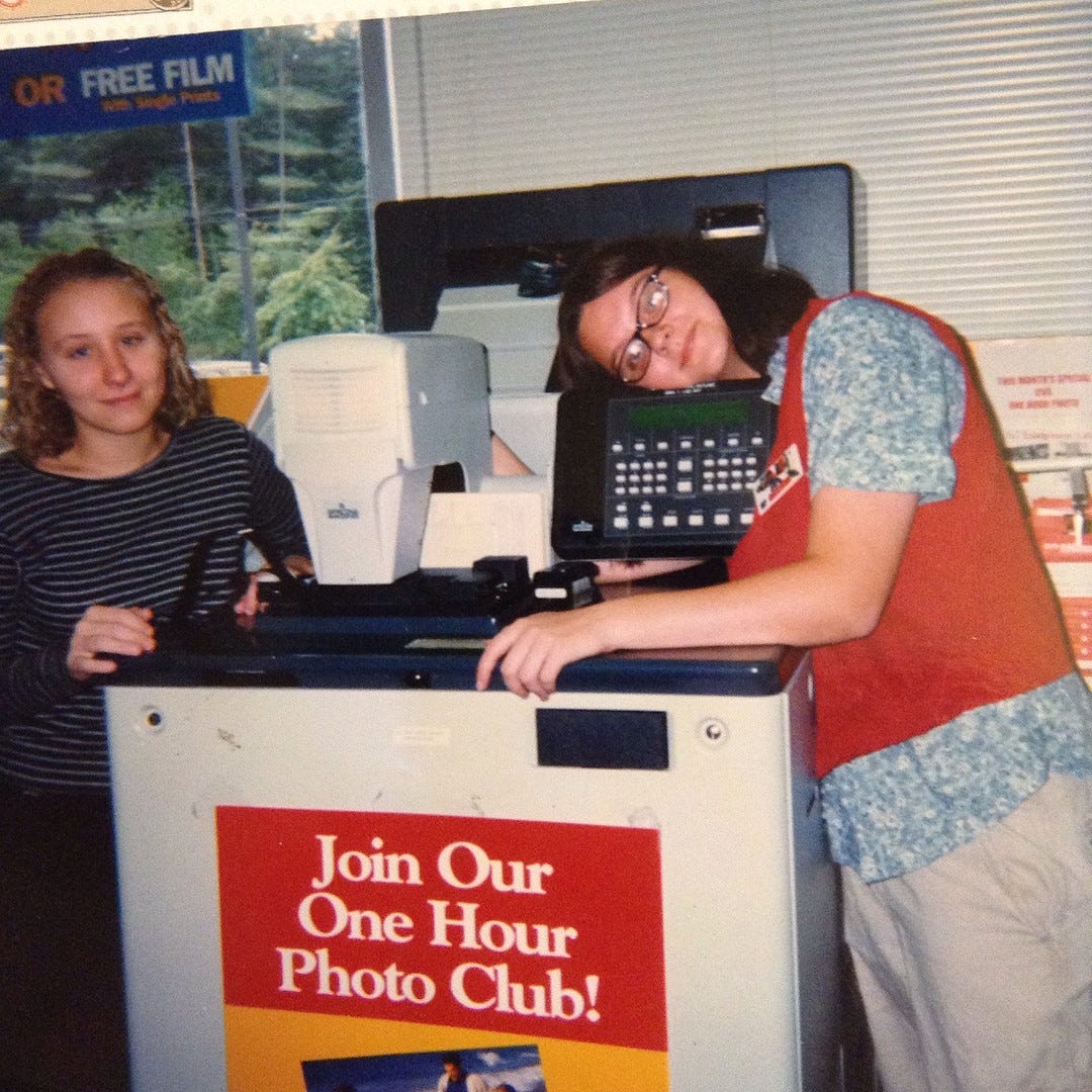 two white teenage girls stand on either side of a 1-hour photo machine in a suburban CVS in the year 1999. They are both wearing late-90s outfits. The author is on the right, with glasses and dark hair, leaning her head on the top of the machine and looking tired.