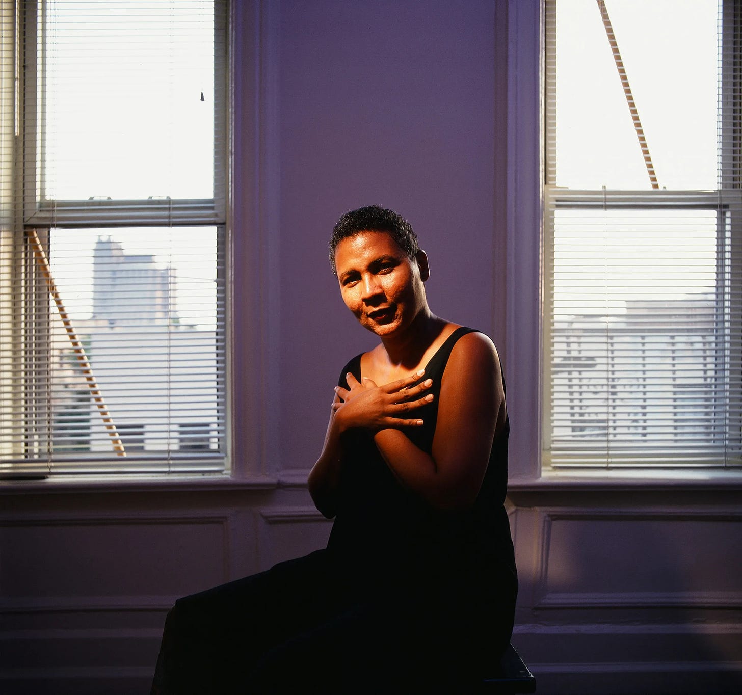 bell hooks is lit by an overhead light as she crosses her hands at her chest and the blue-ish purple light and New York City skyline becomes the background.