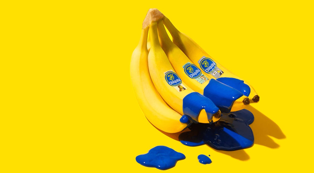 A bunch of bananas with fruit stickers showing famous works of art. There is blue paint splashed on the bananas and they are on a yellow background.