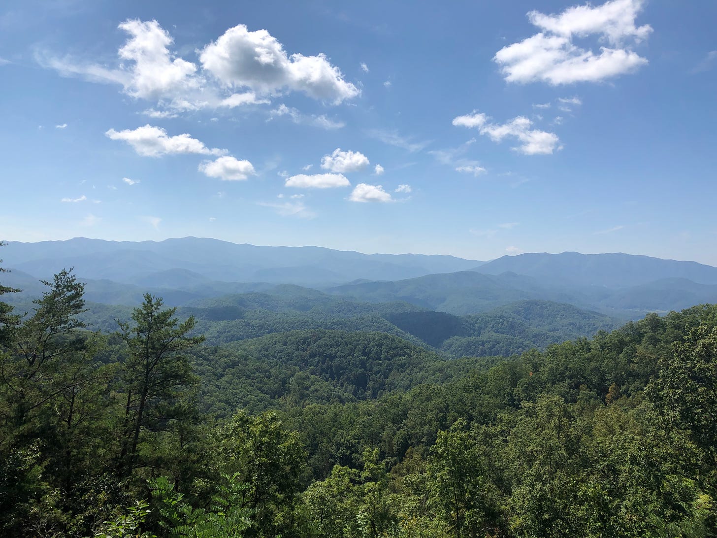 A photo of green mountains stretching into the distance, and blue sky with a few white, puffy clouds