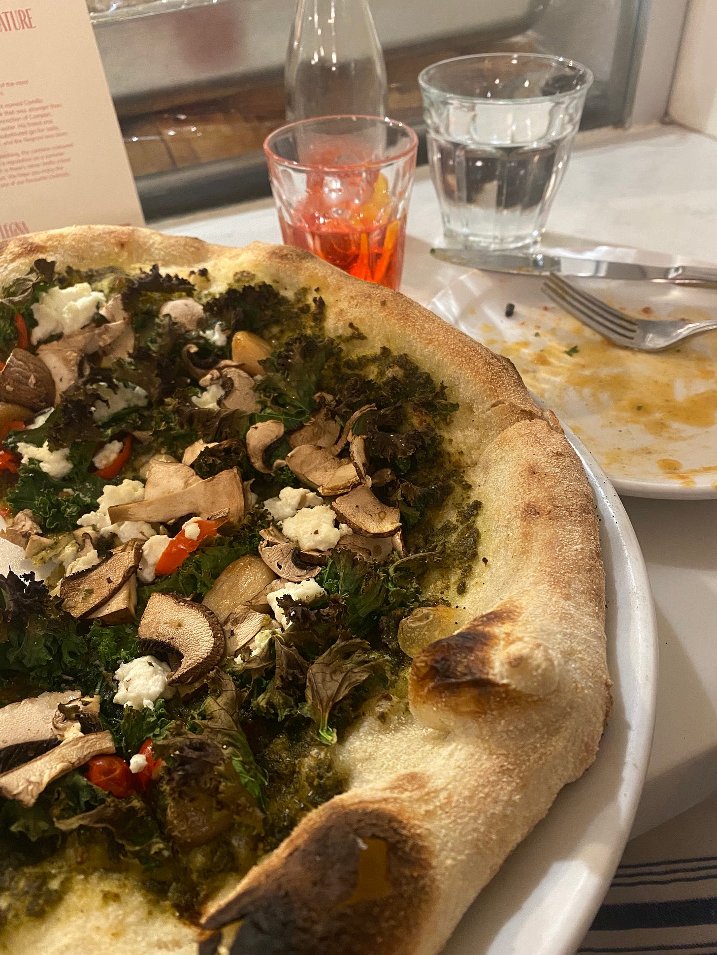 A close-up of the pizza described above, with a saucy-looking empty plate in the background next to a glass of a bicicletta cocktail (the bottle of it is just behind the glass).