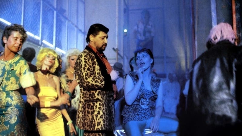 Movie still from Kamikaze '89. Fassbinder stands in a leopard print suit in a club with people dressed in colourful outfits.