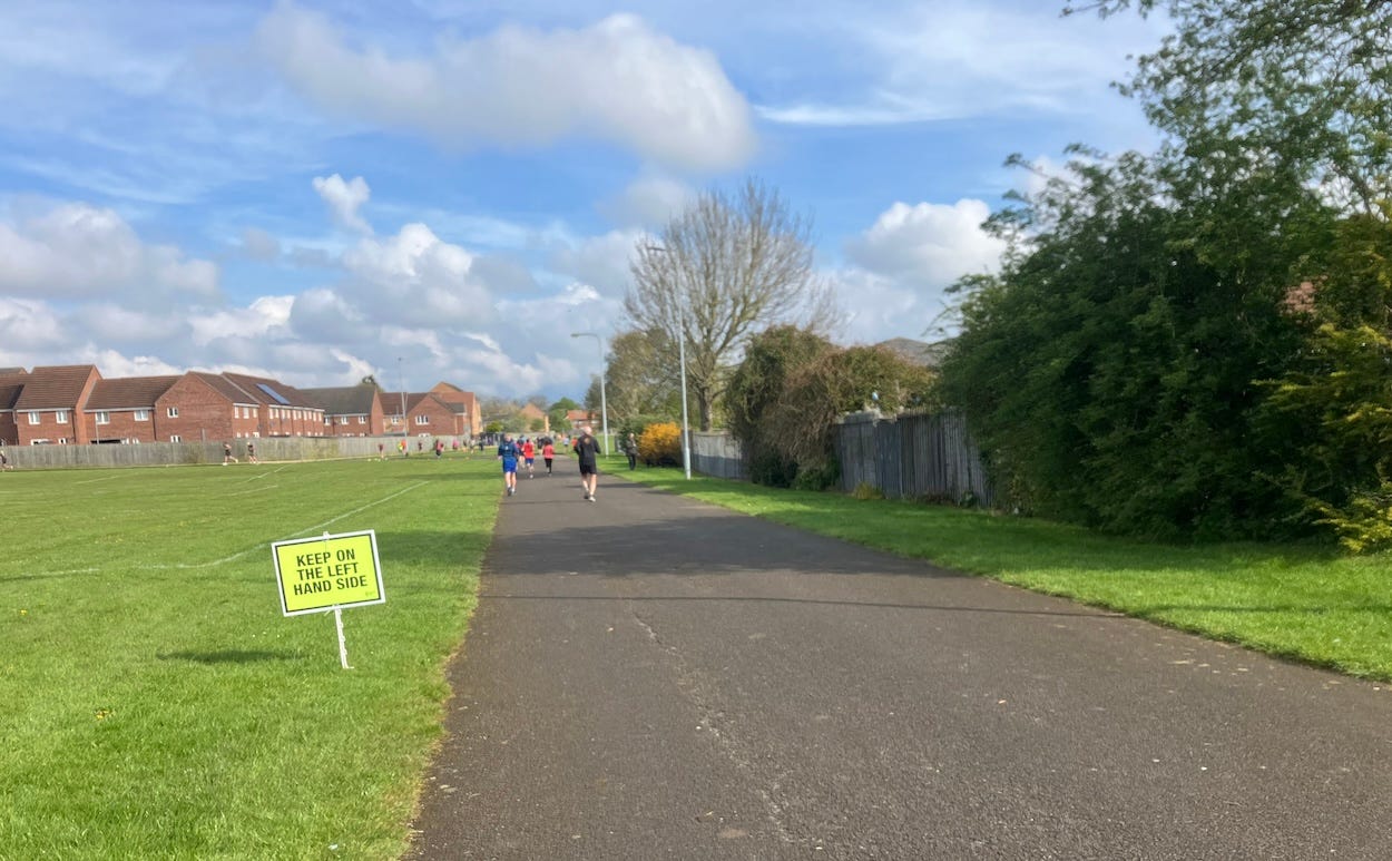 A wide asphalt path leads through a park-like setting with runners in the distance, a keep left sign next to the path, and houses to the right.