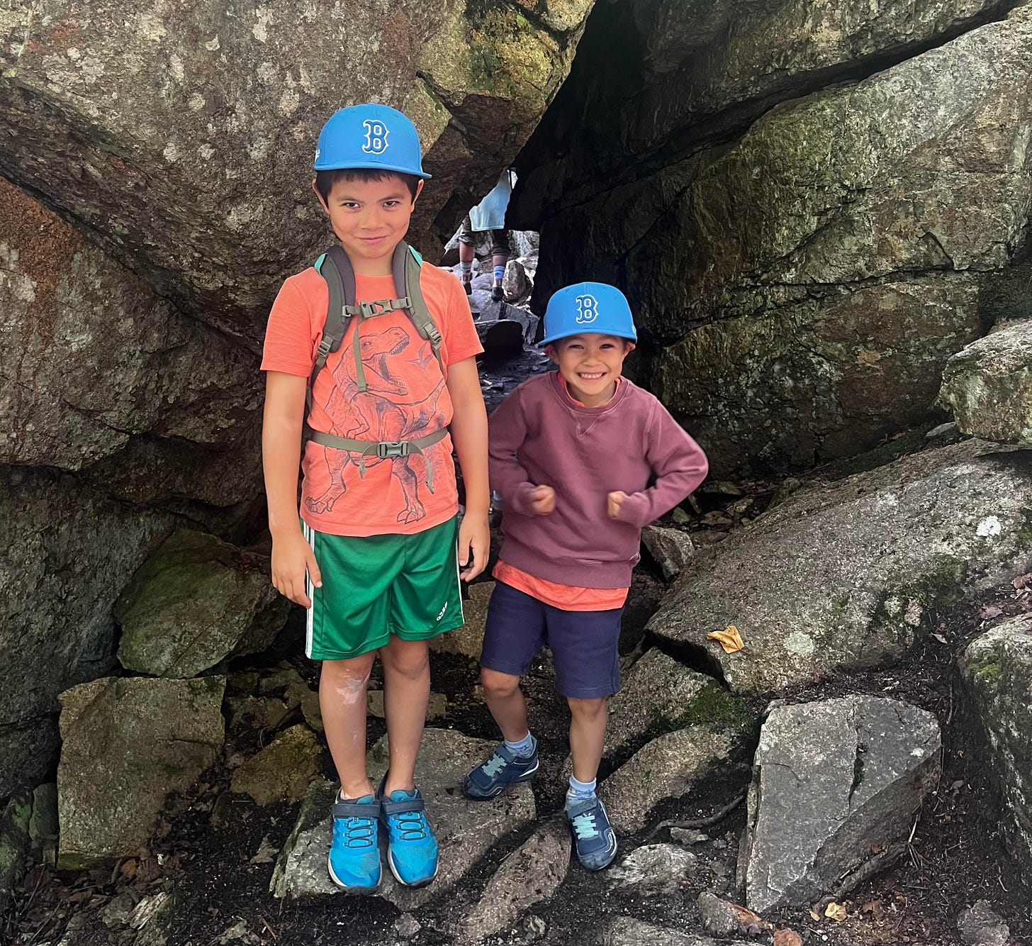 Blaise and Cass pose in front of a tunnel of big boulders wearing blue hats with the Red Sox "B" logo