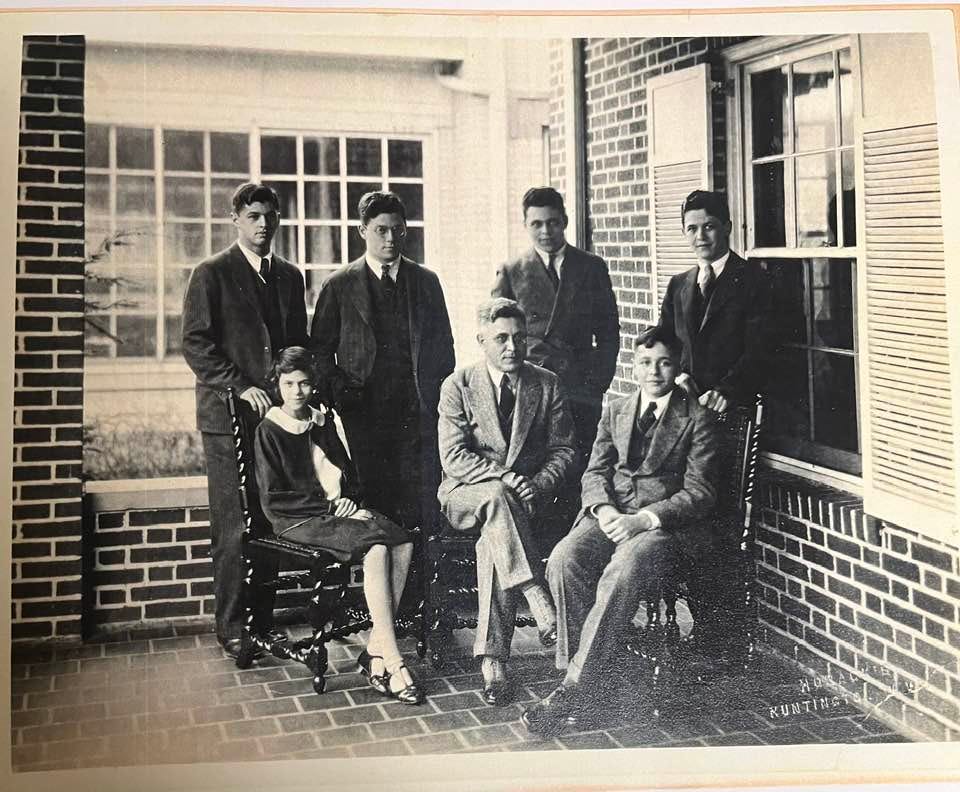 A black and white photo in which an older man in a suit is surrounded by his five sons and one daughter on a porch of a brick house