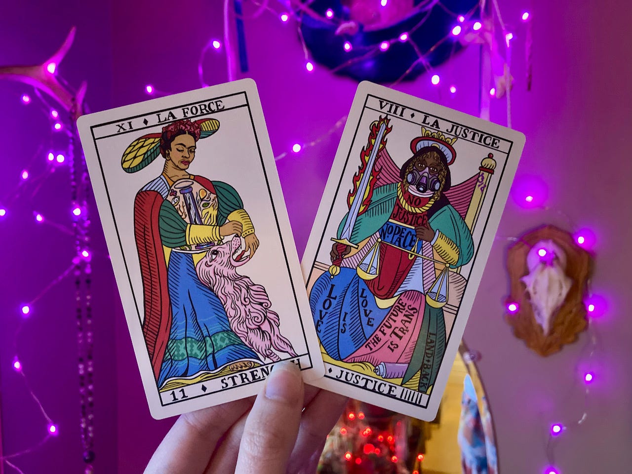 A light-skinned hand holds 2 tarot cards against a background of pink string lights. The Strength card shows Frida Kahlo wearing a painted abdominal cast and gently opening the jaws of a lion. The Justice card shows a dark-skinned person in a gas mask holding a sword and scales, with the slogans "No Justice, No Peace," "Love is Love," "The Future is Trans," and "Land Back" written on their robes.