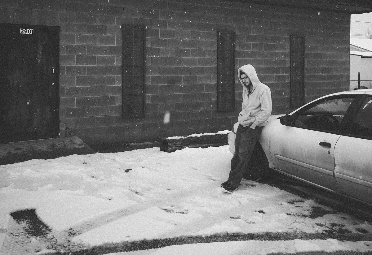 man with glasses and hoodie leaning on car in snowy parking lot