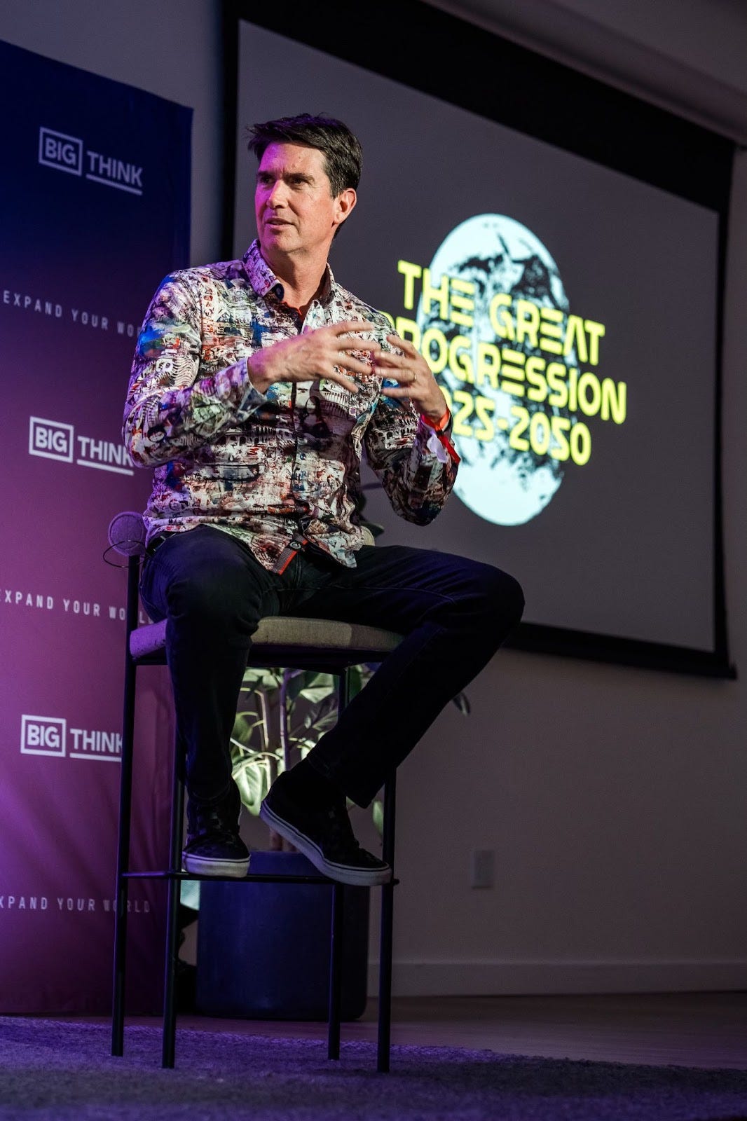 A man sits on a stool speaking, with a screen behind him displaying the text "THE GREAT PROGRESSION 2025-2050" and the Big Think logo visible on banners. A man sits on a stool speaking, with a screen behind him displaying the text "THE GREAT PROGRESSION 2025-2050" and the Big Think logo visible on banners.