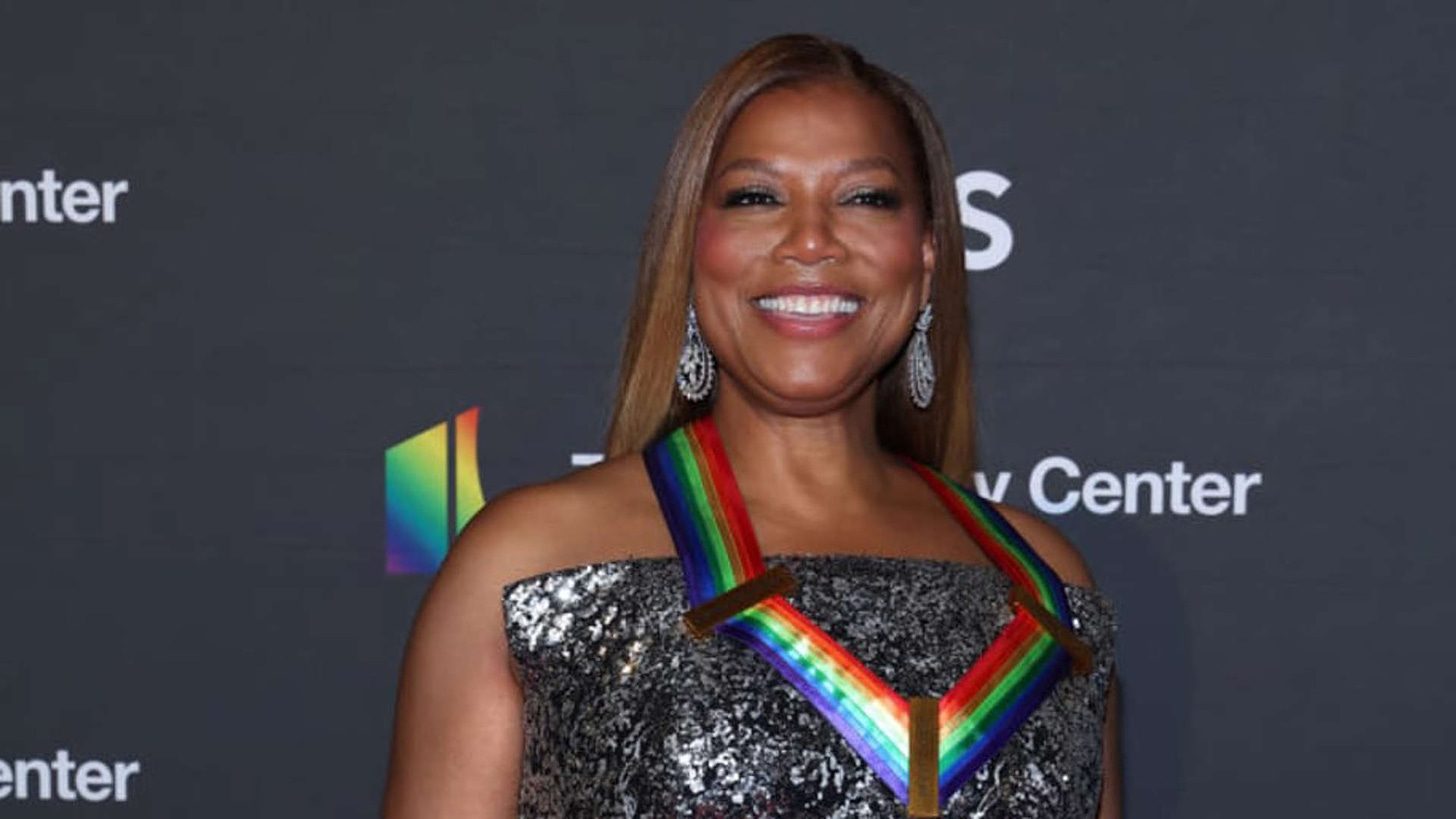 A Black woman with dark blonde hair is smiling broadly while wearing a rainbow Kennedy Center Honoree ribbon over a strapless silver and black dress