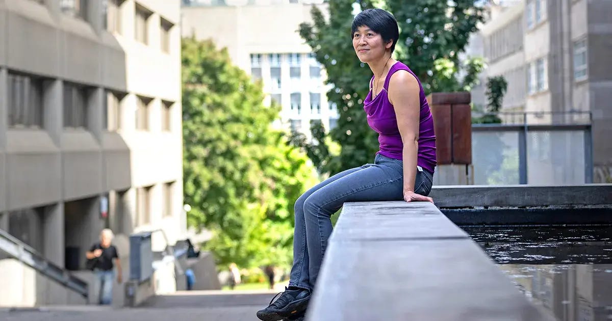 Cynthia Chiang on the McGill University campus. She is perched on a concrete wall, wearing a purple shirt and gray jeans, and the Montreal skyline is in the background.
