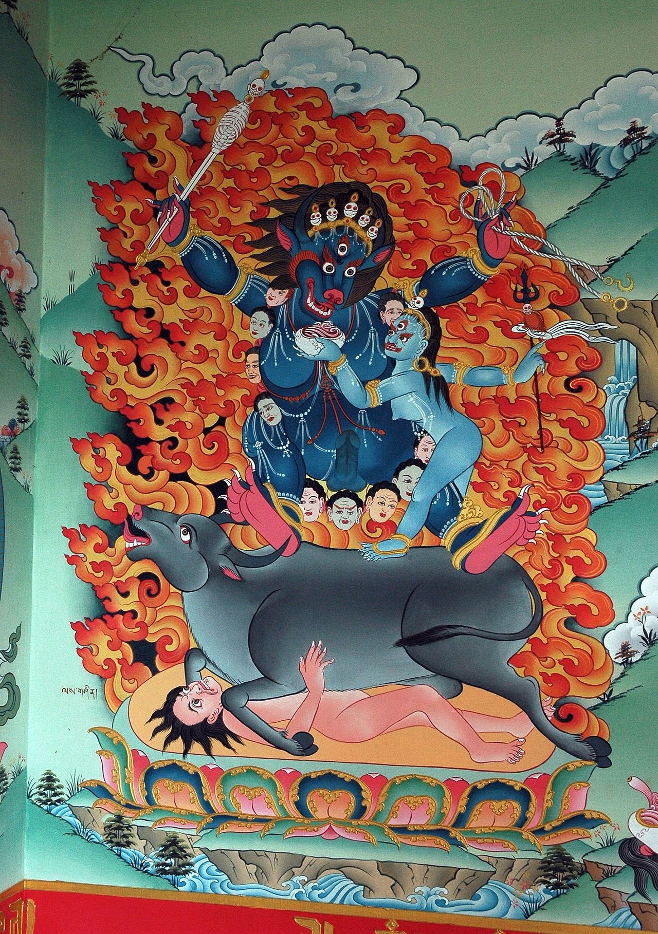 A vibrant and intricate depiction of a deity from Tibetan Buddhist art. The central figure is a fierce, wrathful being with a dark blue body and a ferocious animal-like face, adorned with a crown of skulls and wielding a staff topped with a skeletal figure. Flames radiate dramatically around the deity, symbolizing transformative energy. A secondary, blue-skinned figure assists, holding an object while stepping on a subdued buffalo-like creature. Beneath the buffalo lies a partially clothed human figure in a submissive pose. The background features stylized mountains, trees, and flowing water, characteristic of traditional Tibetan thangka art. The scene conveys themes of power, destruction, and spiritual transformation.