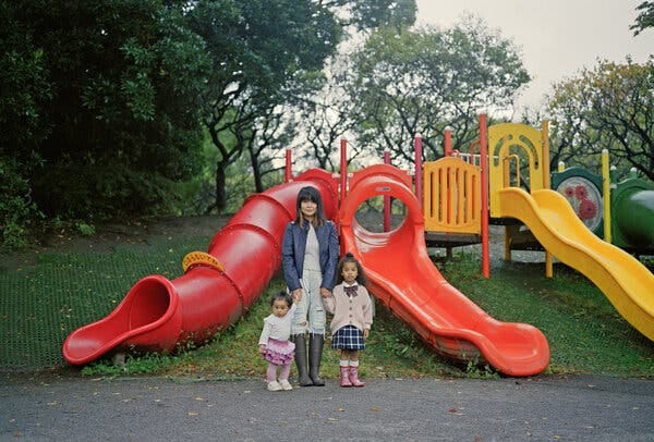 A portrait of a woman holding hands with two small children and standing in front of a jungle gym with slides. 