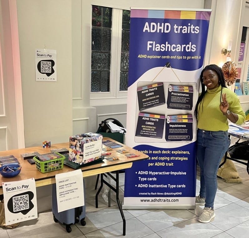 Myself standing by my stall which has sweets, a lucky dip box and the ADHD traits flashcards and a roll banner with pictures and description of the card