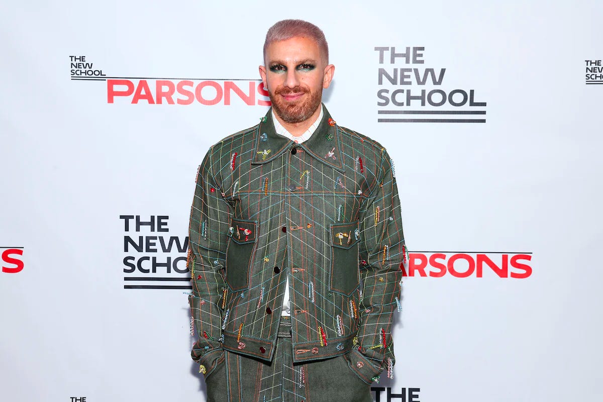 There’s a gay on a step and repeat that has logos for The new School and Parsons. He is wearing an all-denim look, with a green tint and colorful vertical bands of stitches. He’s looking at the camera, smiling, with eye makeup that matches the greens of the garments and dusty lilac hair.