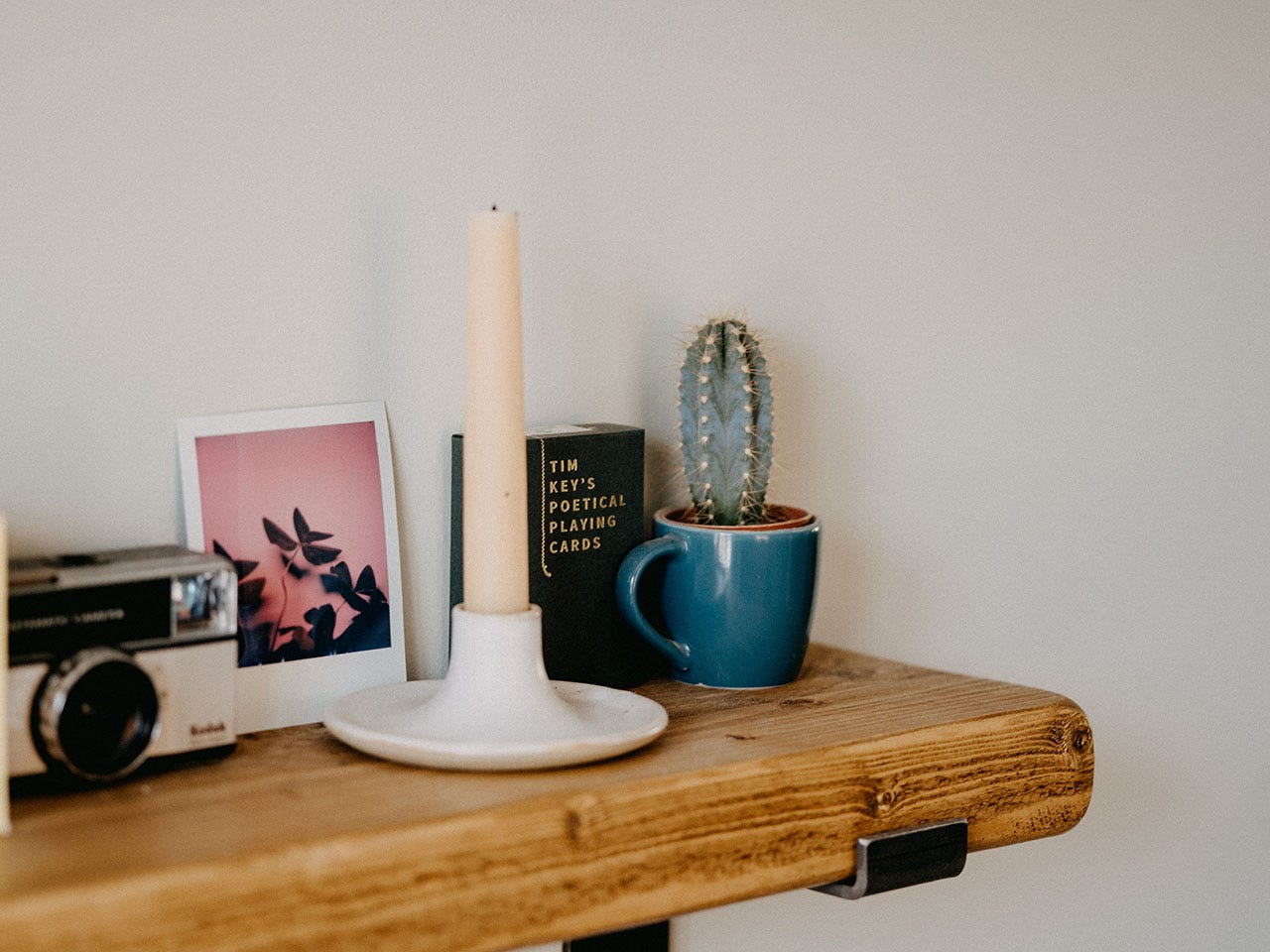 A close-up photo of a wooden bookshelf, on which sits a camera, a polaroid photo of a plant, a candle, a cactus in a blue mug, and 'Tim Key's Poetical Playing Cards'. Photo by Annie Spratt, via Unsplash