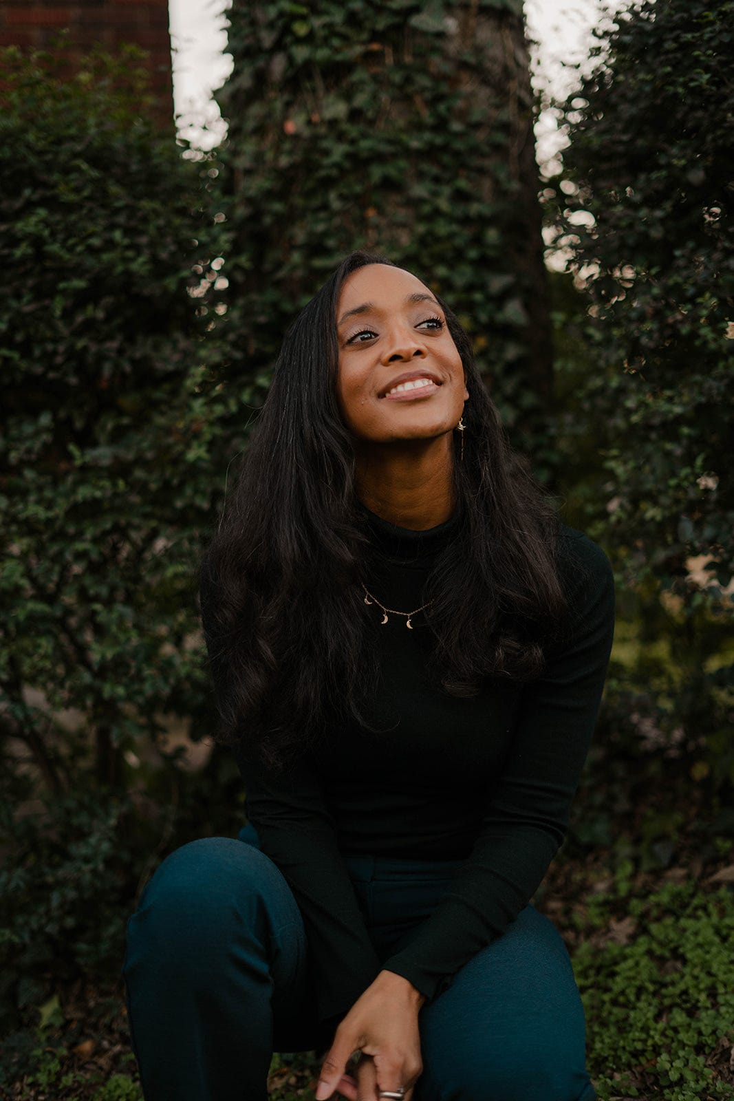 mage description: Black woman kneels above ground in front of large tree trunks covered with green leaves. Woman is wearing Black turtle neck and blue pants. She looks toward the sky in a hopeful manner.