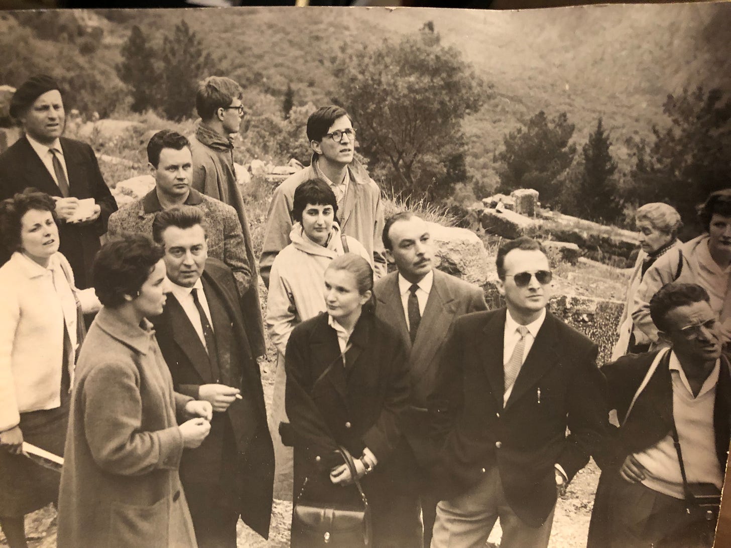 A group of 15 adults from the 1950s are standing on a hillside with a ruin behind them, talking with one another, their eyes directed above at something. One woman looks sideways at the others.
