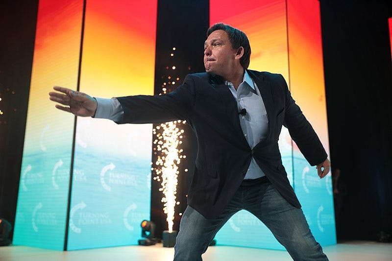 Ron DeSantis, his lips pursed and right arm arm outstretched dramatically, leaning forward a bit as if casting a spell. Behind him, a small onstage prop spits a thin fountain of sparks