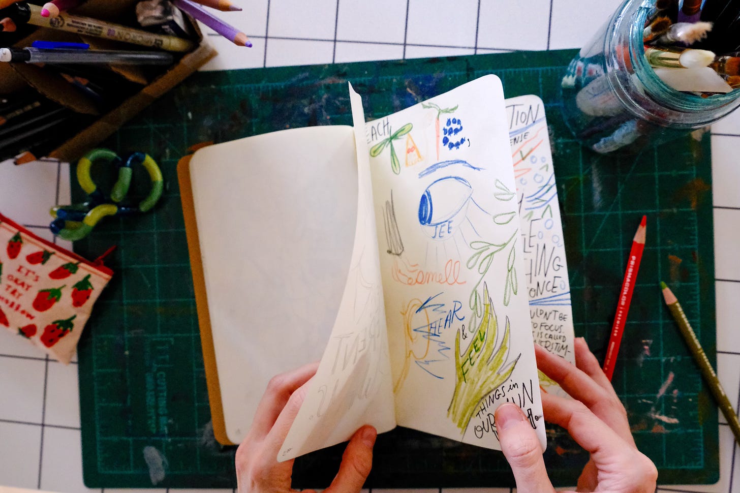 Gracie flipping through a sketchbook with sensory sketches. Pencils and a green tangle surround workspace.