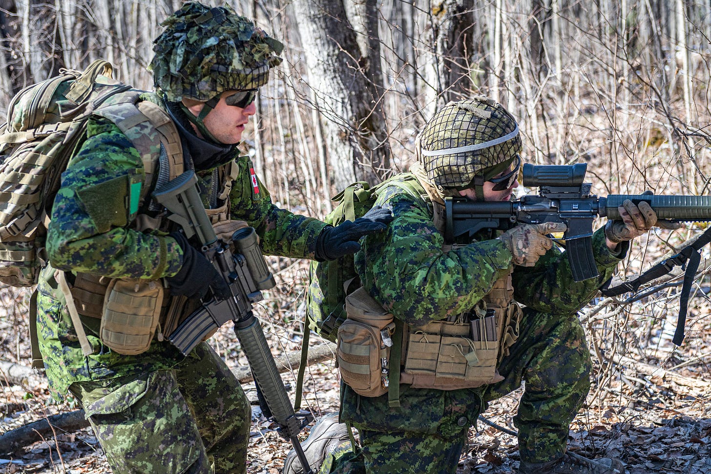 One soldier kneels on the ground aiming his weapon, while another stands behind him in a sunny, wooded area.