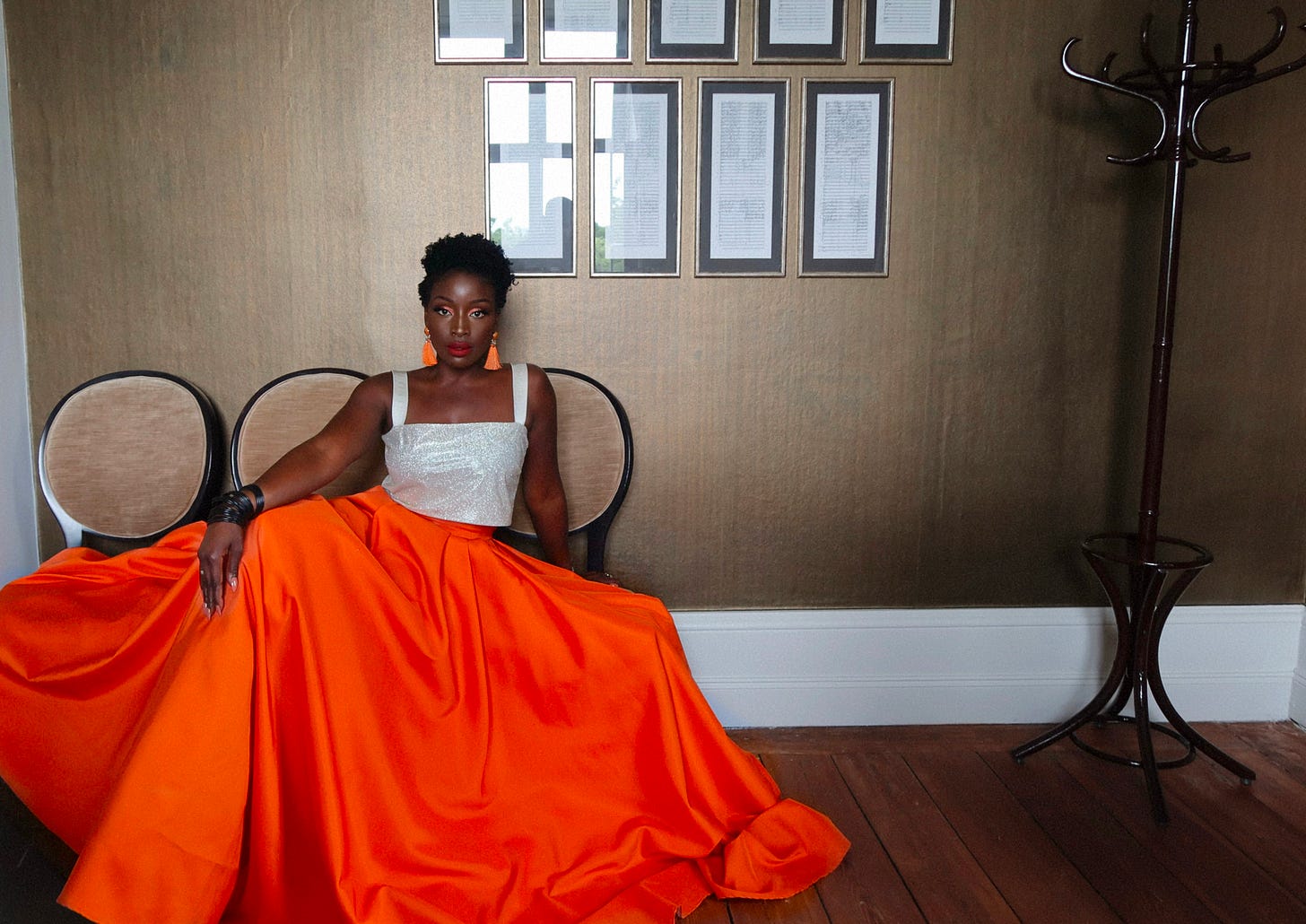 Lovette Jallow seated in an orange gown, looking sideways in a moment of introspection, surrounded by framed documents.