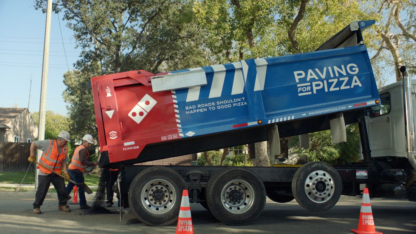 An industrial dumptrick branded with Dominos Pizza branding dumps asphalt over a pothole